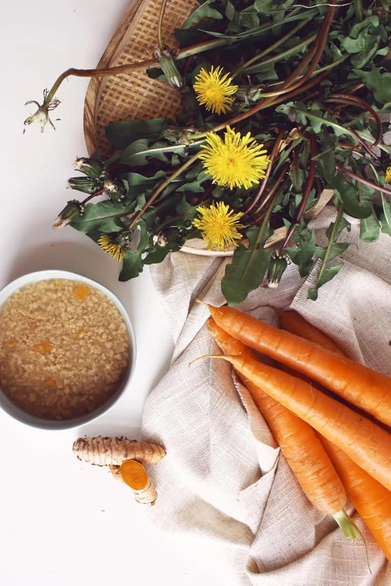 Schale mit Hühnersuppe, daneben Karotten, Kurkuma und Löwenzahnblüten